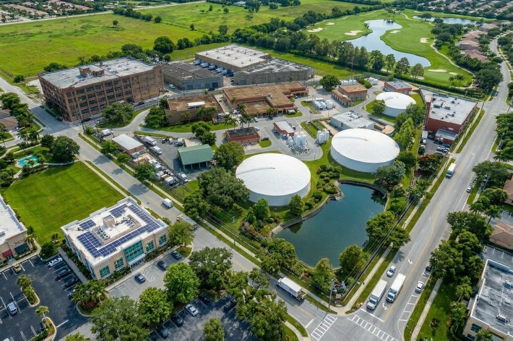 Aerial view of a utility facility with large storage tanks, surrounded by industrial buildings, parking areas, and nearby residential development.